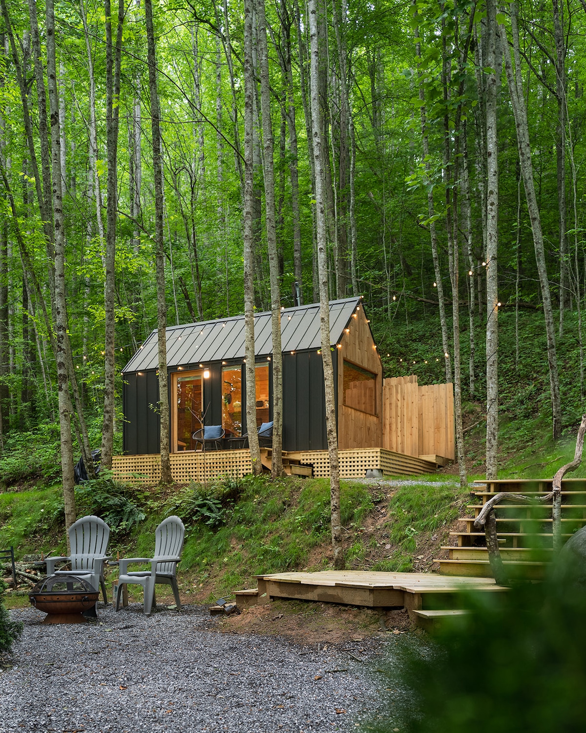 A Nordic-style tiny house is set amidst lush greenery, featuring wood and glass elements. A welcoming deck leads to a private entrance, surrounded by tall trees. Outdoor seating is provided near a small fire pit, inviting relaxation in a serene natural environment.