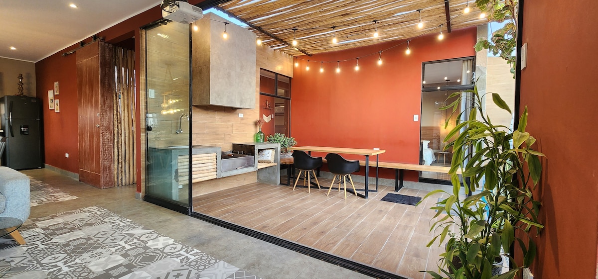 An inviting open space is showcased, featuring a dining area with a large wooden table and black chairs. A warm orange wall complements the modern kitchen visible in the background. Large glass doors provide a seamless transition to the outdoor area, enhanced by soft overhead lighting.