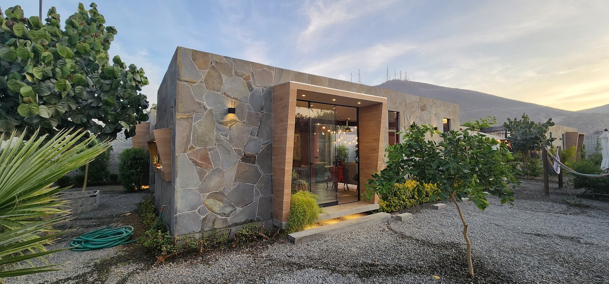 A spacious house constructed with a combination of stone and wood materials is framed by lush greenery. The entrance features large glass doors, inviting natural light into the interior. Various plants surround the structure, enhancing the serene outdoor environment.
