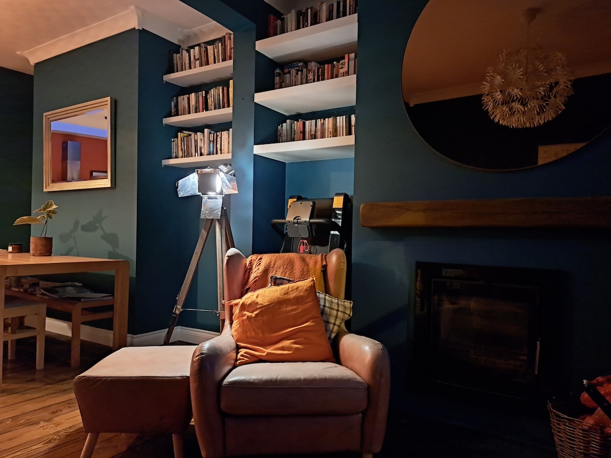 A cozy corner is presented with a comfortable armchair adorned with cushions and a matching ottoman. A sleek lamp brightens the area, while bookshelves filled with an array of books add character to the space. A round mirror reflects light, enhancing the ambiance.