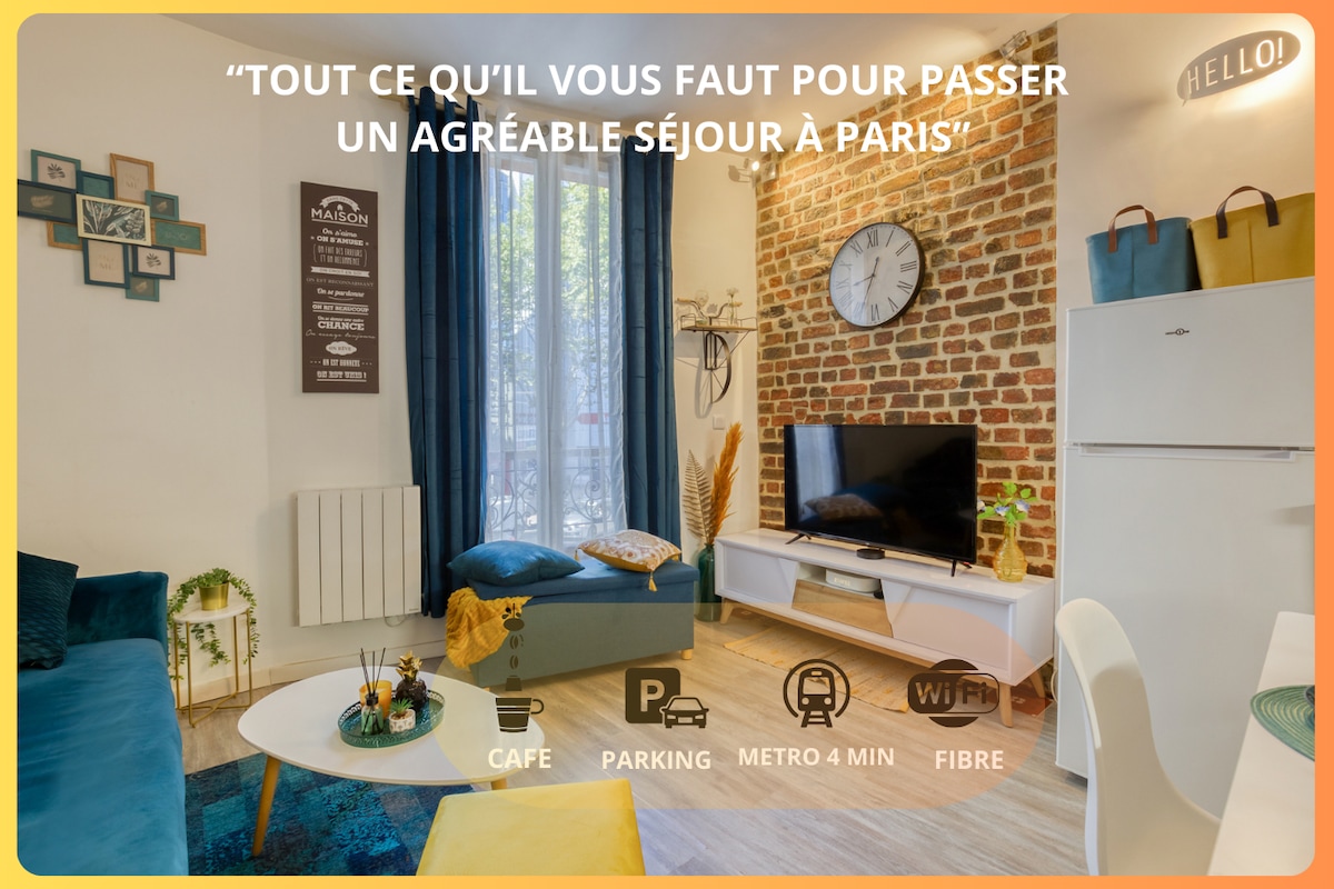A bright living area features a mix of modern and rustic decor, highlighted by a brick accent wall and large windows dressed in blue curtains. A comfortable blue sofa and a small table are arranged for relaxation. A refrigerator and a small kitchenette are visible in the space.