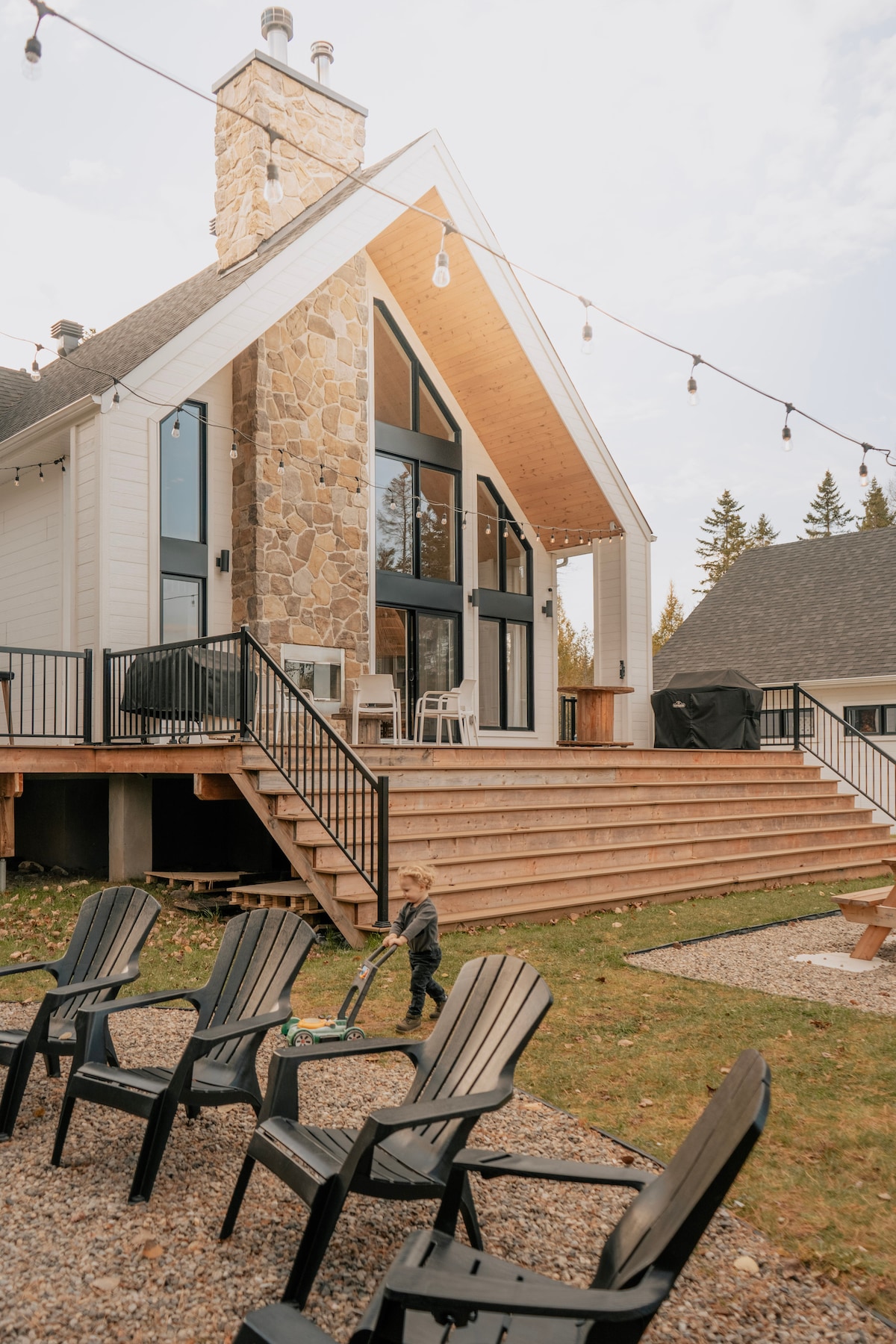 The exterior of the chalet features a modern farmhouse design with a large stone chimney and expansive windows. A wooden deck extends from the house, leading to a spacious outdoor area with several black chairs. Warm lighting from string lights creates a welcoming ambiance.