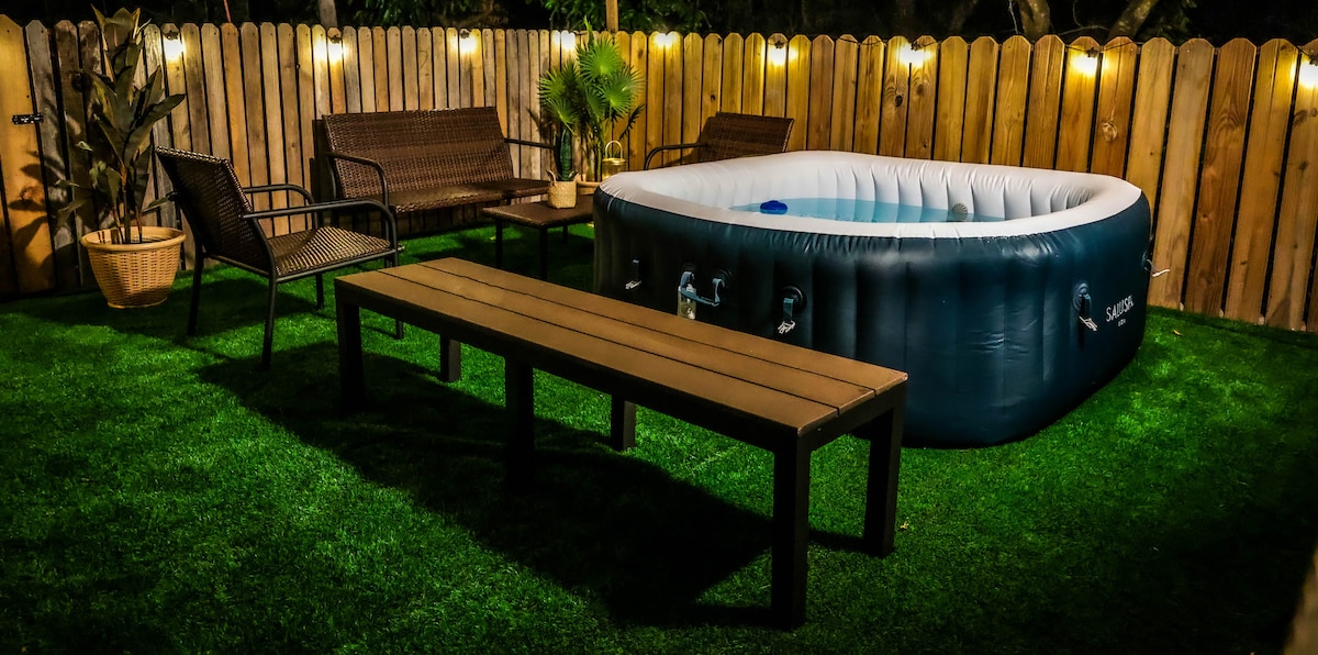 An outdoor patio is featured, adorned with lush green grass. A blue hot tub sits prominently, surrounded by brown wicker seating and a rectangular table. Soft lighting illuminates the area, creating a tranquil setting for relaxation and gatherings.