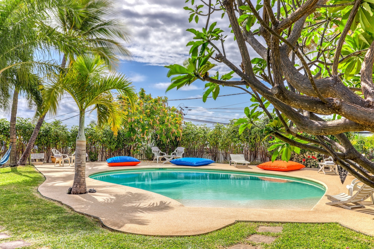 A curvilinear swimming pool is surrounded by a tropical garden. Lounge chairs and colorful bean bags are placed nearby, providing seating options. Palm trees offer shade, while vibrant greenery adds to the serene landscape under a partially cloudy sky.