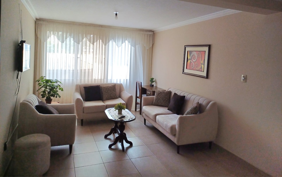 A cozy living area features two light-colored sofas with decorative cushions, arranged around a wooden coffee table. Natural light filters through sheer curtains, illuminating the space. A small dining table is placed in the corner, accompanied by a potted plant adding a touch of greenery.