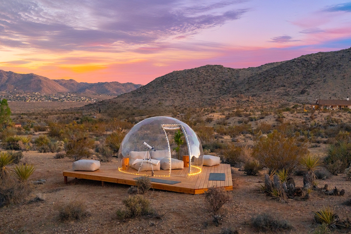 A transparent dome is nestled within the desert landscape, illuminated by soft lights. Surrounding the dome are cushions arranged on a wooden deck. The backdrop features rolling hills and a vibrant sunset sky with hues of pink and purple.