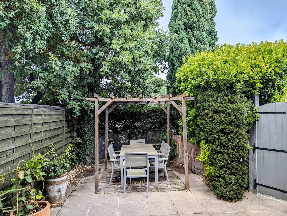 A shaded outdoor patio area features a wooden pergola supporting climbing plants. A large rectangular table is surrounded by several chairs, providing a tranquil space for gatherings. Lush greenery from trees and shrubs creates a serene atmosphere, with decorative planters positioned along the patio edges.