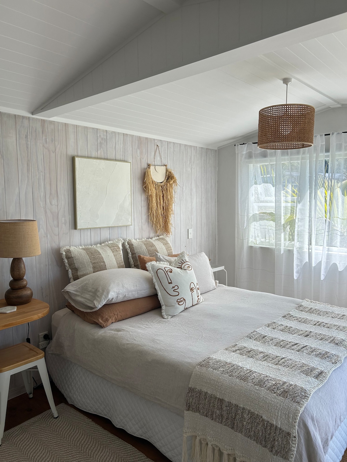 A serene bedroom features a spacious bed adorned with textured pillows and a neutral-colored blanket. Natural light filters through a sheer curtain, illuminating the warm timber accents and soft furnishings. A nightstand with a lamp complements the calming decor.