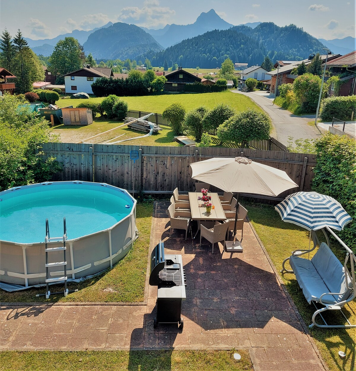 An outdoor area is presented, featuring a circular above-ground pool adjacent to a patio set with a table and chairs. A striped umbrella provides shade, while a grill is positioned nearby. Lush greenery and distant mountains create a scenic backdrop.