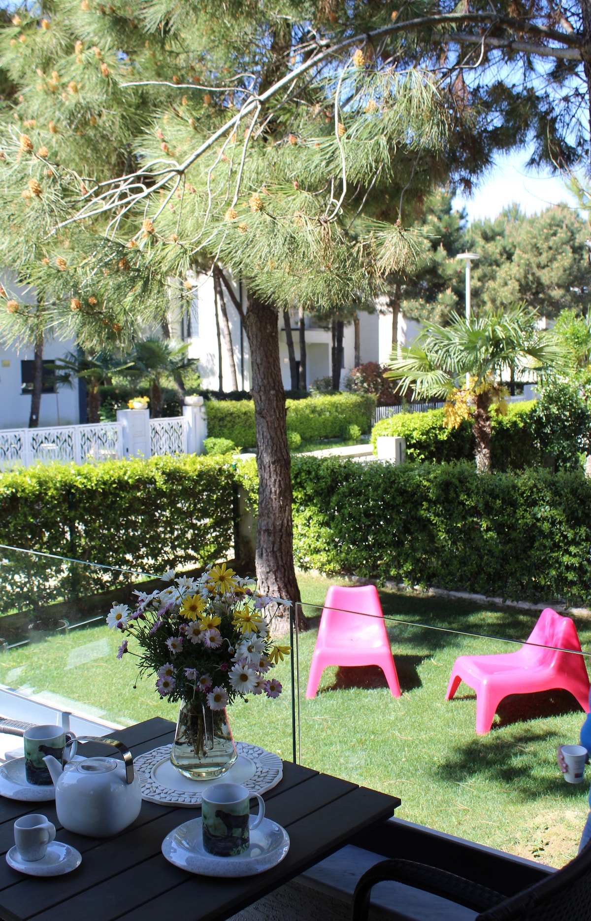 A patio table is adorned with a vase of flowers, tea set, and cups, overlooking a lush green garden. Two pink chairs are positioned on the grass, framed by well-maintained hedges and trees, creating a serene outdoor space.