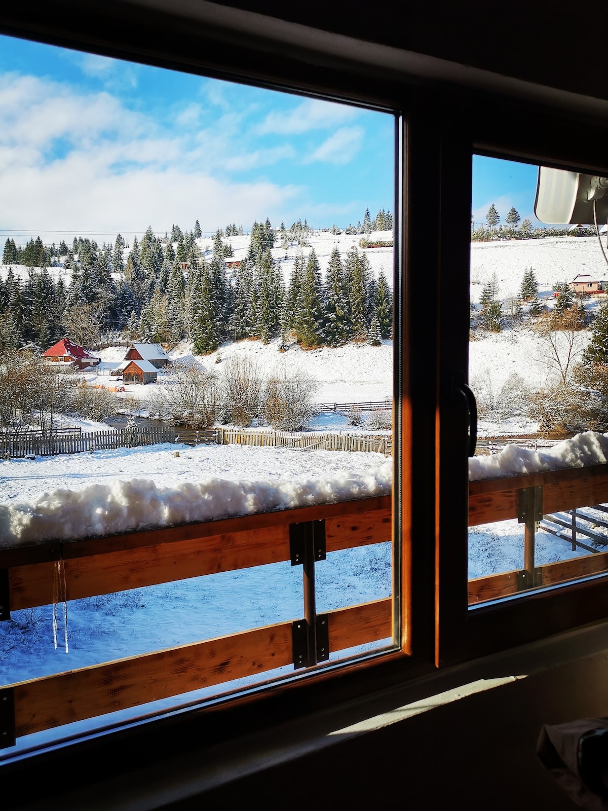 A view from a large window displays a snowy landscape, featuring a mix of pine trees and wooden houses. The balcony railing is adorned with a layer of fresh snow, complementing the serene winter scene outside, under a partly cloudy sky.