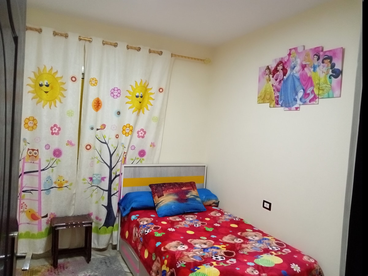 A bright bedroom features a bed with vibrant bedding and character designs. Colorful curtains adorned with sun and flower patterns frame a window. A small wooden stool stands near the bed, while a wall art piece displays classic animated characters.