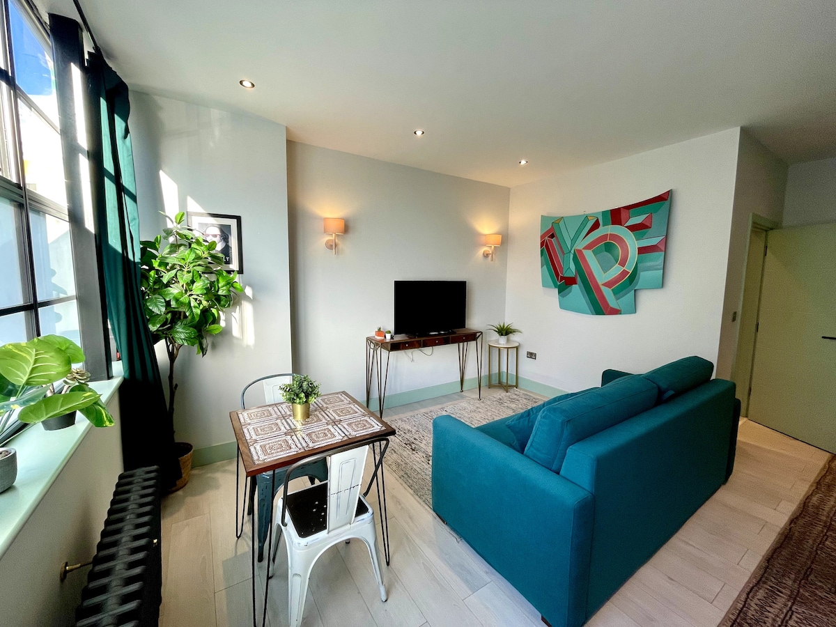 A cozy lounge area features a spacious teal sofa facing a wall-mounted television. A small dining table with white metal chairs sits nearby, adorned with a potted plant. The room is illuminated by natural light from large windows, enhanced by contemporary wall art.