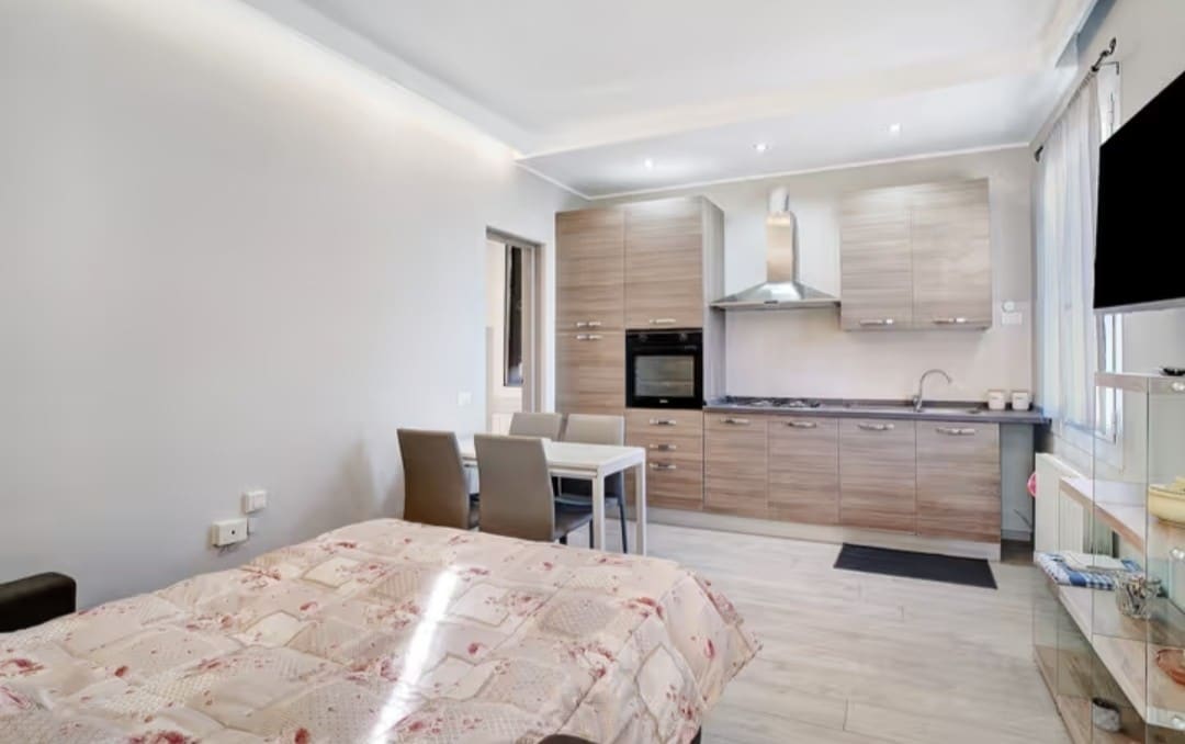 A modern kitchenette is featured with wooden cabinetry and stainless steel appliances. A dining table with four chairs is positioned near the window. A comfortable sofa bed is visible in the foreground, accompanied by a light quilt and natural light illuminating the inviting space.