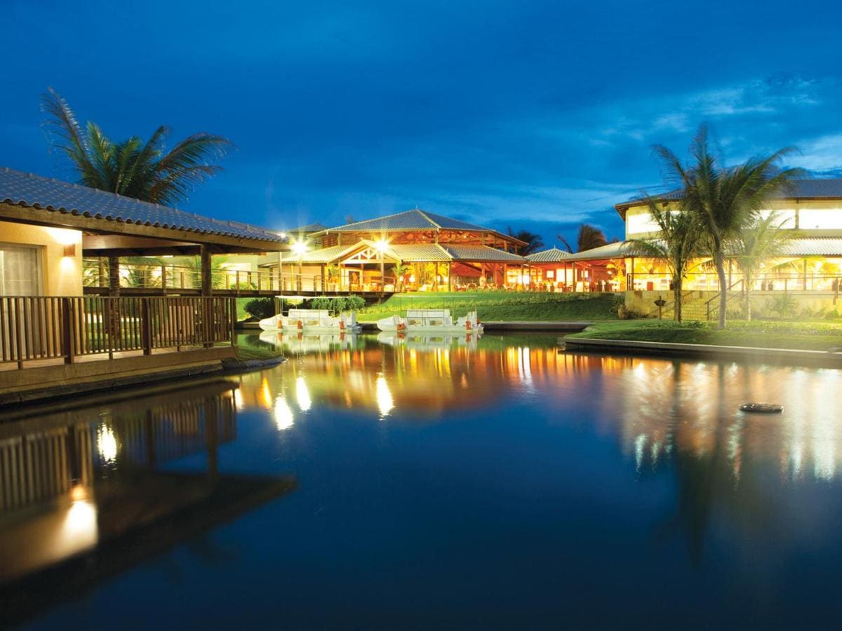 The resort's elegant exterior is reflected in the tranquil waters of a nearby pond, illuminated by soft lighting. Surrounded by palm trees, the dining area and lounge spaces are visible, creating a serene atmosphere for relaxation and enjoyment.