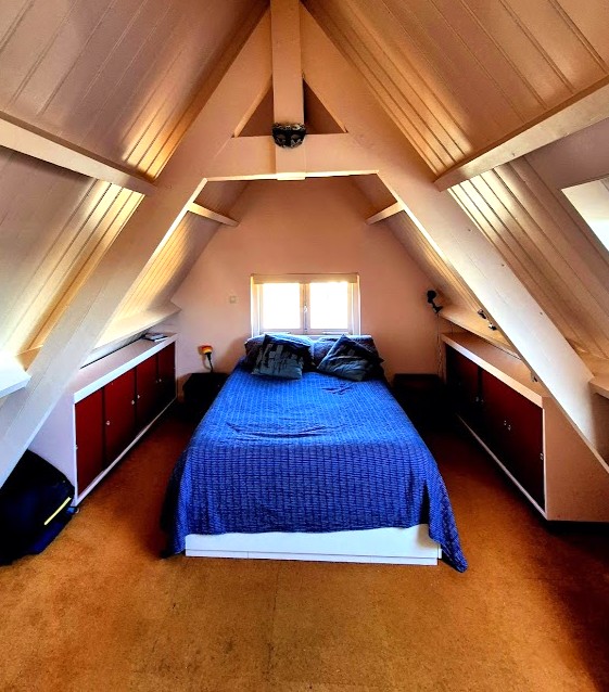 A private bedroom features a sloped ceiling with exposed wooden beams. A double bed is dressed in blue bedding, flanked by built-in storage cabinets. Natural light filters in through a small window, illuminating the clean lines and cozy atmosphere of the space.