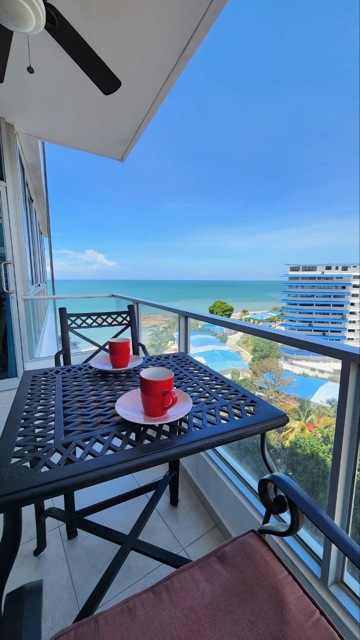 A small outdoor balcony is furnished with a black metal table and two matching chairs. Two red mugs sit atop the table, offering a view of the ocean and distant buildings. The sky is clear with light clouds, reflecting a serene coastal atmosphere.