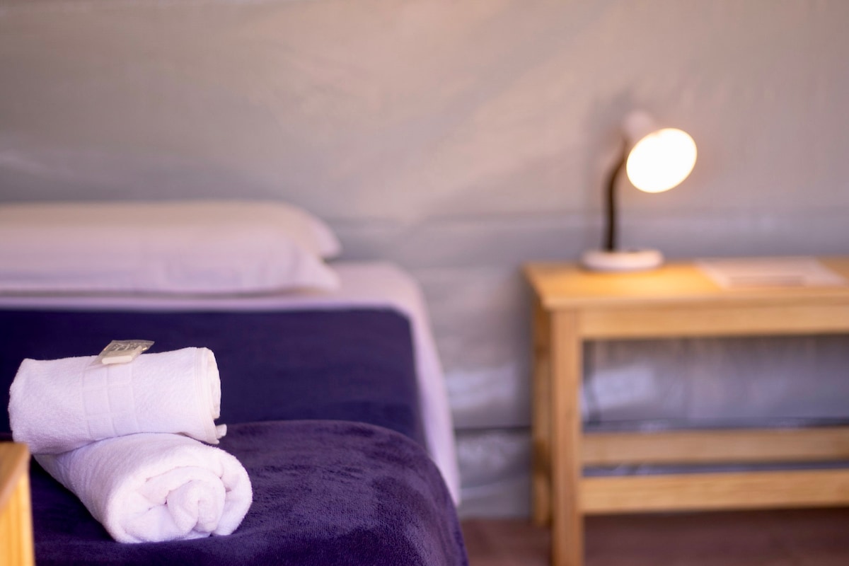 A cozy glamping space features a comfortable bed with soft pillows. A neatly rolled towel is positioned on a blanket, while a lamp on a wooden side table provides warm illumination. The interior showcases simple, natural materials against a neutral backdrop.
