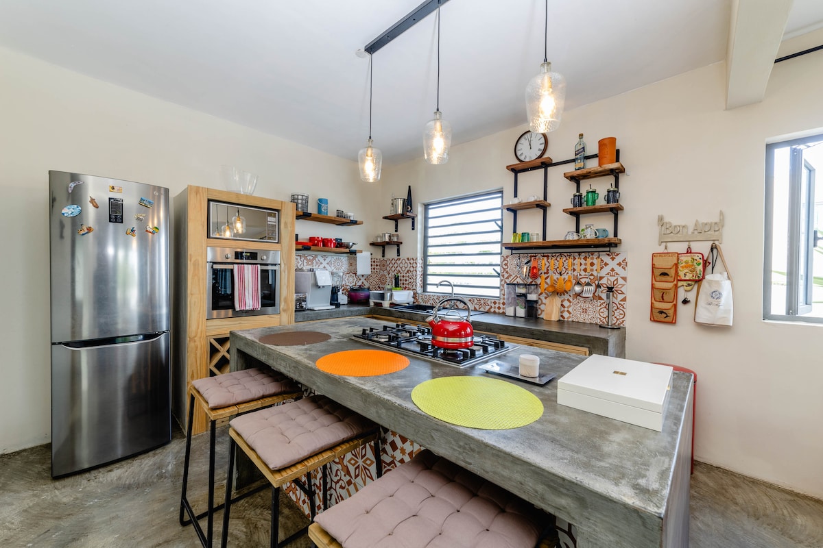 A contemporary kitchen features a large concrete island with seating, complemented by colorful placemats. Open shelving displays various kitchen items, and modern appliances, including a stainless steel refrigerator and an oven, are visible. Natural light enters through window openings, enhancing the space.