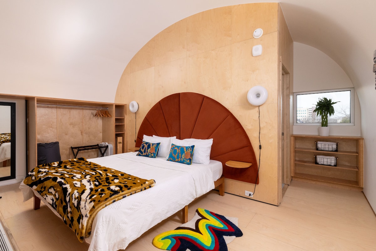 A lofted bedroom is centered around a spacious bed featuring a unique headboard with a curved design. Natural light fills the room, highlighting wood paneling and minimalist shelving. A colorful rug adds a vibrant touch to the neutral decor, while a window provides additional views.