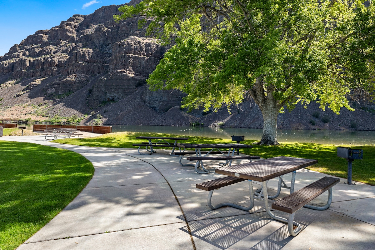 A serene outdoor space is depicted, featuring several picnic tables arranged along a paved path. Lush green grass provides a comfortable area, while a large tree offers shade. Rocky cliffs and a calm body of water form the backdrop, enhancing the natural setting.