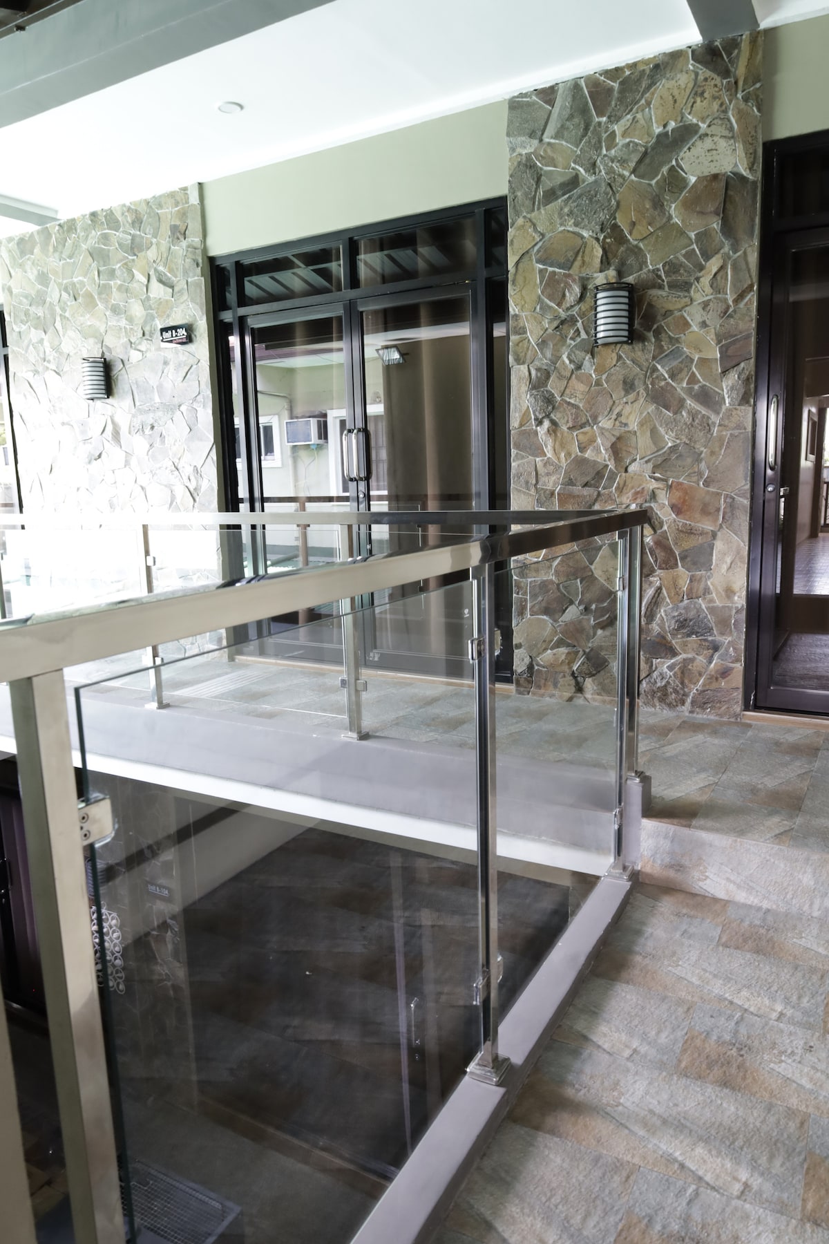 A well-structured entrance features a combination of stone walls and glass railings. A concrete pathway leads to double glass doors, inviting guests into the interior. Soft lighting fixtures mounted on the stone wall add visibility, highlighting the modern design.