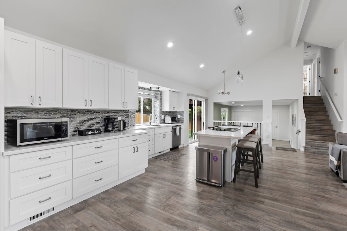A modern kitchen is showcased with sleek white cabinetry and a spacious island. Appliances including a microwave, coffee maker, and utensils are visible. Ample counter space facilitates meal preparation, while large windows bring in natural light, illuminating the wood-like flooring throughout the area.