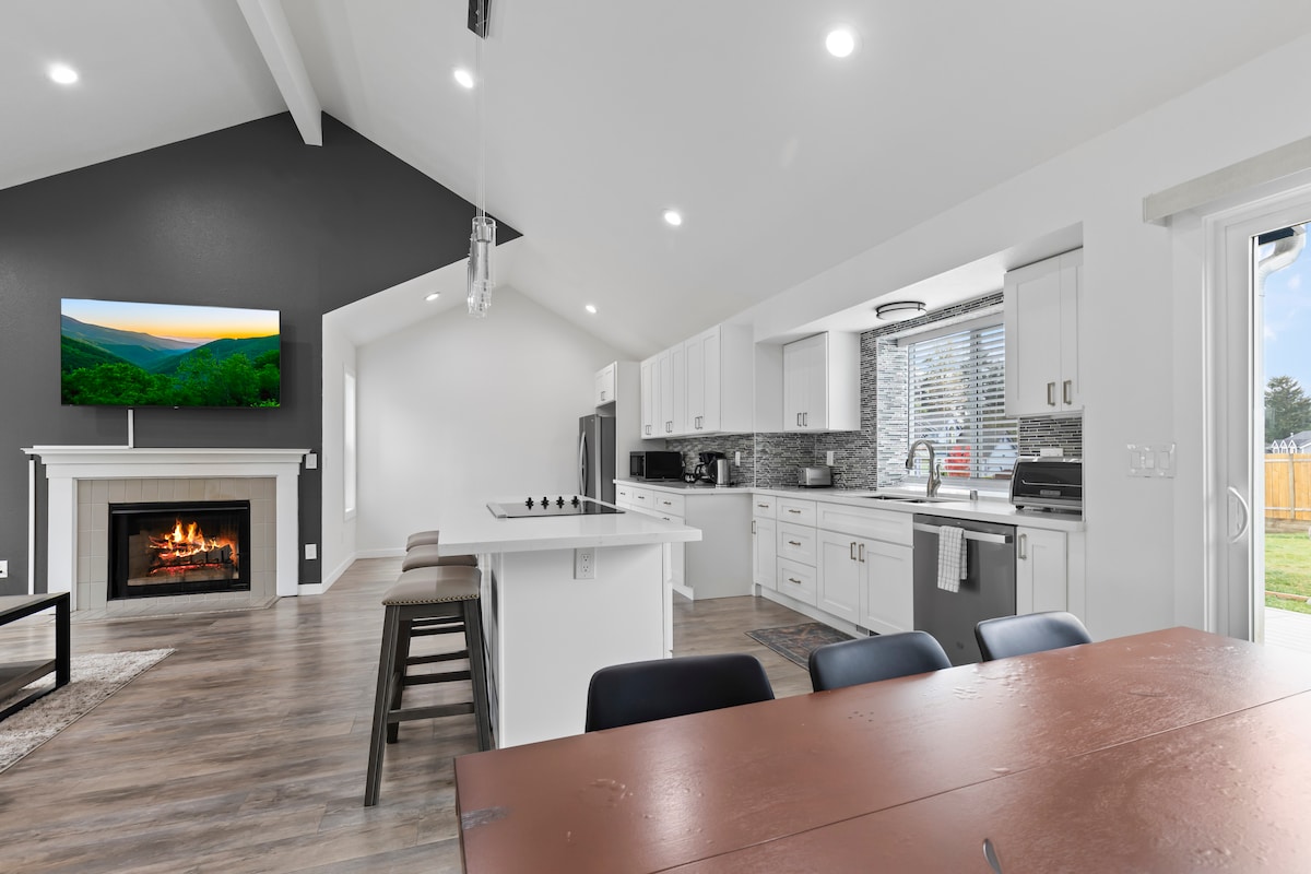 The kitchen area features modern appliances and a large island with seating for four. A cozy living space includes a fireplace and a smart TV mounted on the wall, while natural light streams through the windows, enhancing the open layout.