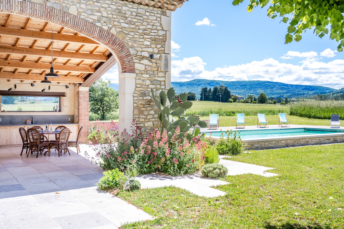 A spacious outdoor area includes a covered terrace with a dining table and rattan chairs. The scene features a refreshing pool surrounded by sun loungers, with scenic views of the surrounding fields and hills in the background. Lush garden plants add natural beauty to the space.