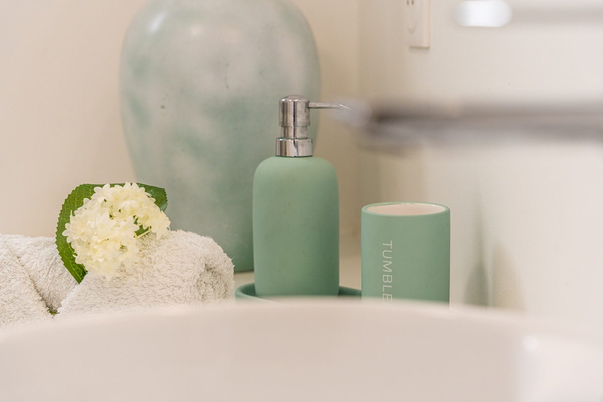 A neatly arranged bathroom counter is displayed, featuring a soft white towel and a delicate flower. A mint green soap dispenser and a cup labeled 'TUMBLER' add a calming touch, complemented by a light blue decorative vase in the background.