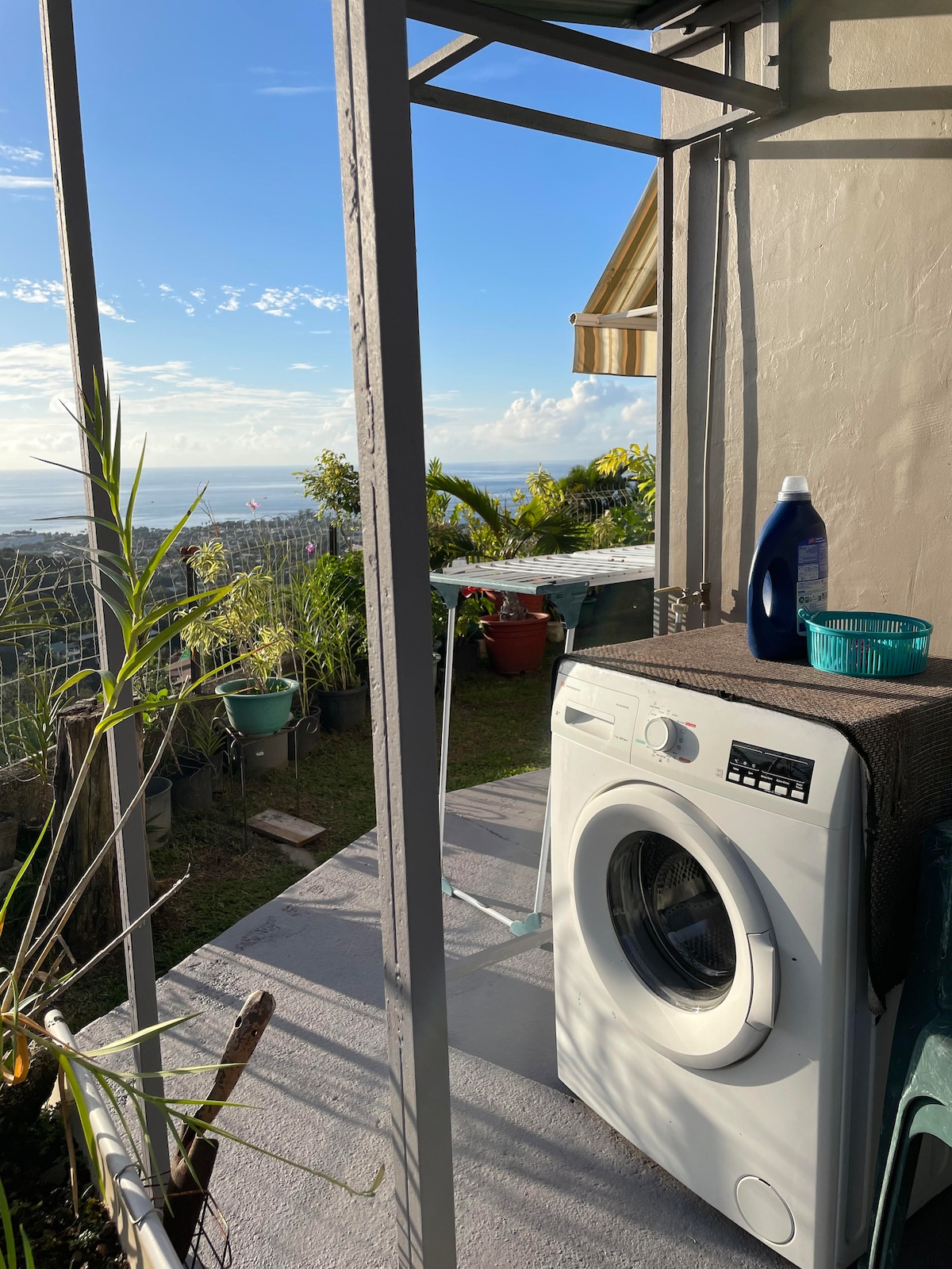 A washing machine is positioned on a terrace, surrounded by greenery. Scenic views of the landscape and ocean are visible in the background, complemented by a clear sky. An outdoor area with a small table is included, enhancing the space for relaxation.