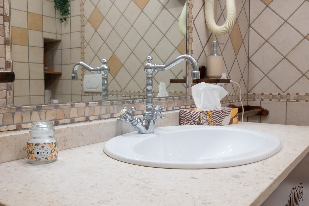 A spacious bathroom features a large mirrored wall above a white, circular sink. A vintage-style faucet is positioned to the side, complemented by a small decorative candle and a box of tissues nearby. Neutral tiles create a soothing ambiance throughout the space.