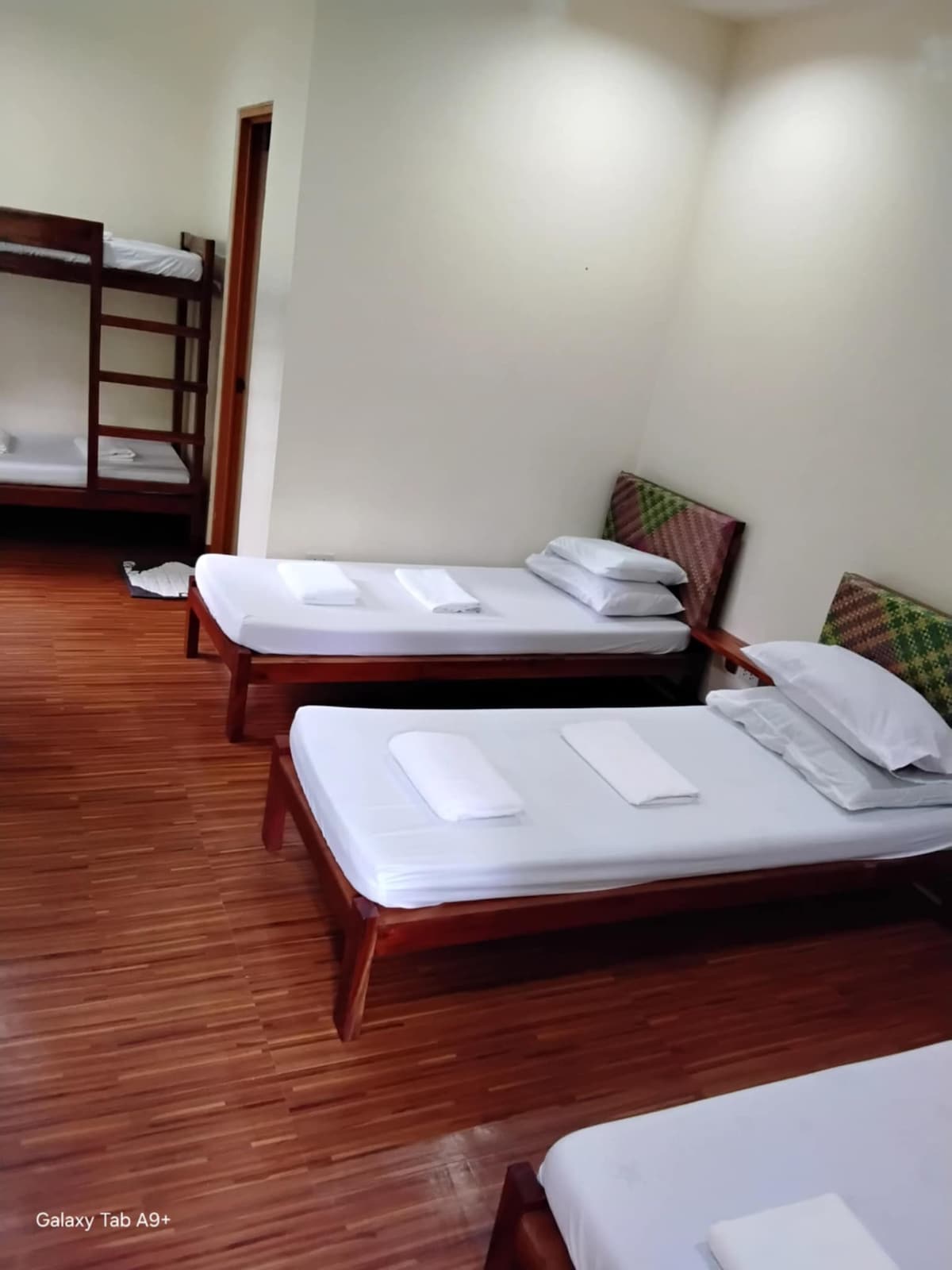 The room contains three beds arranged in a spacious layout, featuring wooden frames with clean white linens. A set of bunk beds is positioned in the corner, enhancing sleeping options for guests. Natural light fills the room from a nearby window.