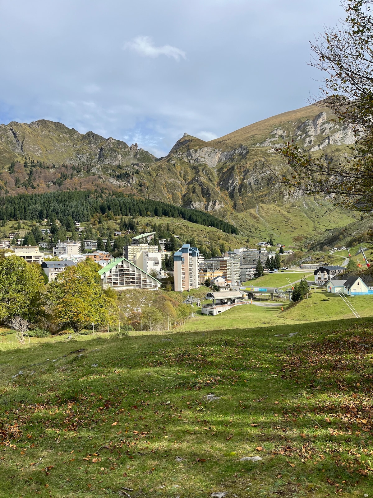A view showcases the surrounding mountains and valley, with various buildings nestled among trees. The landscape is marked by slopes and greenery, highlighting the natural beauty of the area. The sky is partly cloudy, enhancing the mountainous backdrop.