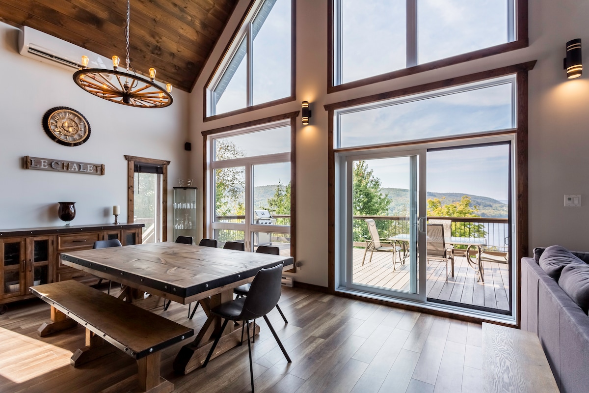 A bright and spacious living area includes large glass doors that offer scenic views. Two main seating areas are visible: a long wooden dining table surrounded by chairs and a comfortable sofa. Natural light enhances the wooden accents of the cabin's interior.