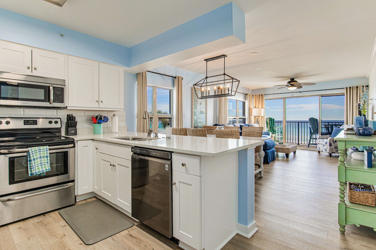 A fully equipped kitchen features modern stainless steel appliances, including an oven and dishwasher. White cabinetry contrasts with a light blue wall. A spacious island with seating is visible, while sunlight filters into the open living space, showcasing a panoramic Gulf view through large windows.