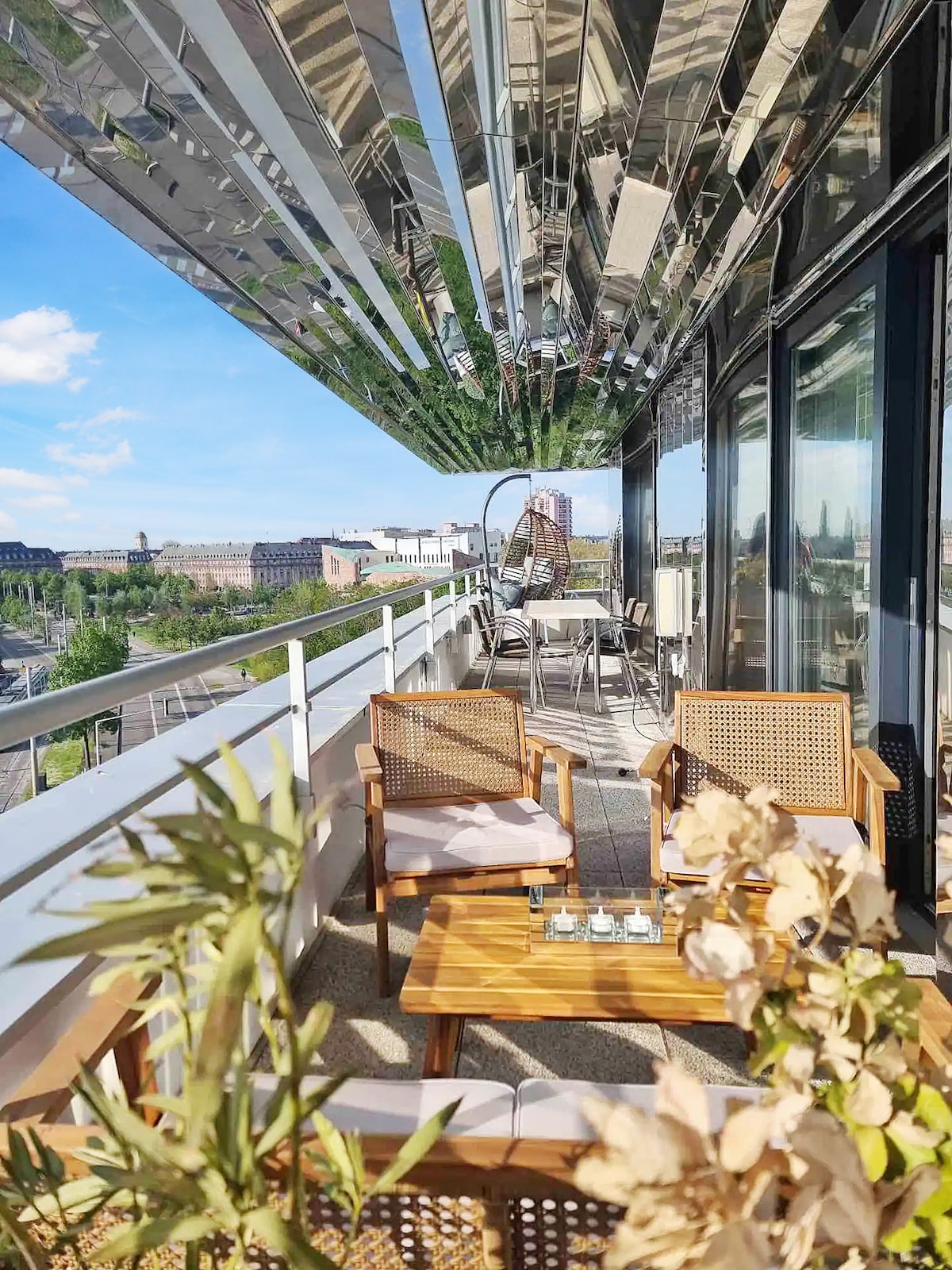 An outdoor terrace is furnished with a wooden table and two woven chairs. A reflective canopy provides shade, while plants frame the view. The cityscape of Strasbourg can be seen in the background, showcasing a blend of modern and historical architecture.