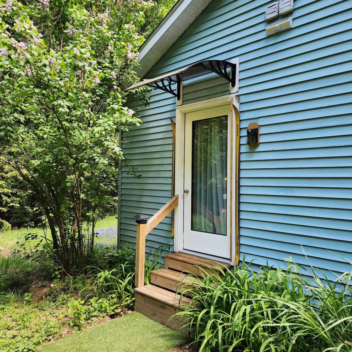 A private entrance is visible, featuring white double doors under an overhead awning. The entrance is flanked by lush greenery and floral landscaping. A set of three wooden steps leads up to the doorway, providing access to the rental space.