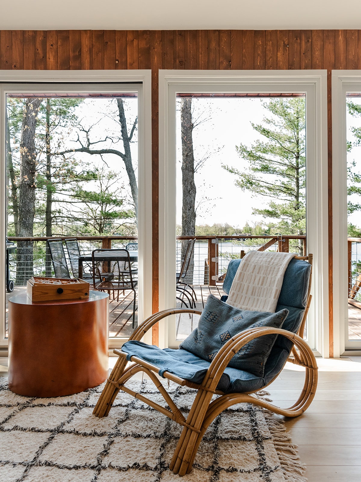 A comfortable chair is positioned near large windows, allowing for views of the deck and surrounding trees. A small side table is nearby, while a patterned rug complements the setting. Natural light fills the space, enhancing the serene ambiance.