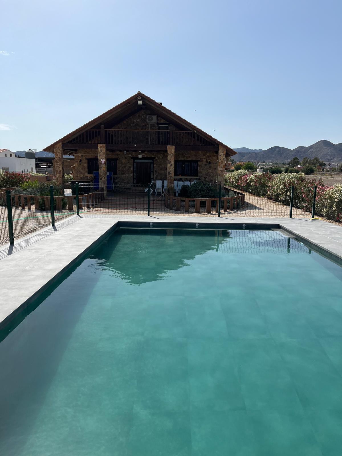 A tranquil swimming pool is situated in the foreground, reflecting the clear sky. The rustic stone house is visible, featuring a covered porch and a wooden balcony. Lush greenery and mountains provide a scenic backdrop, enhancing the serene environment.