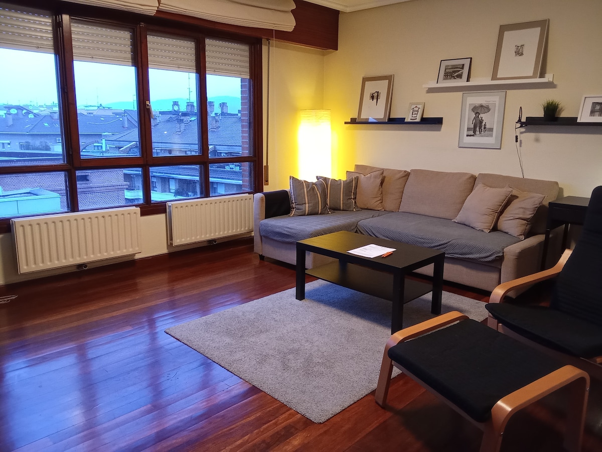 The living area features a spacious sofa positioned adjacent to a large window, allowing ample natural light. A low table sits on a soft rug, complemented by a comfortable chair. Shelves displaying framed artwork add a personal touch to the inviting space.