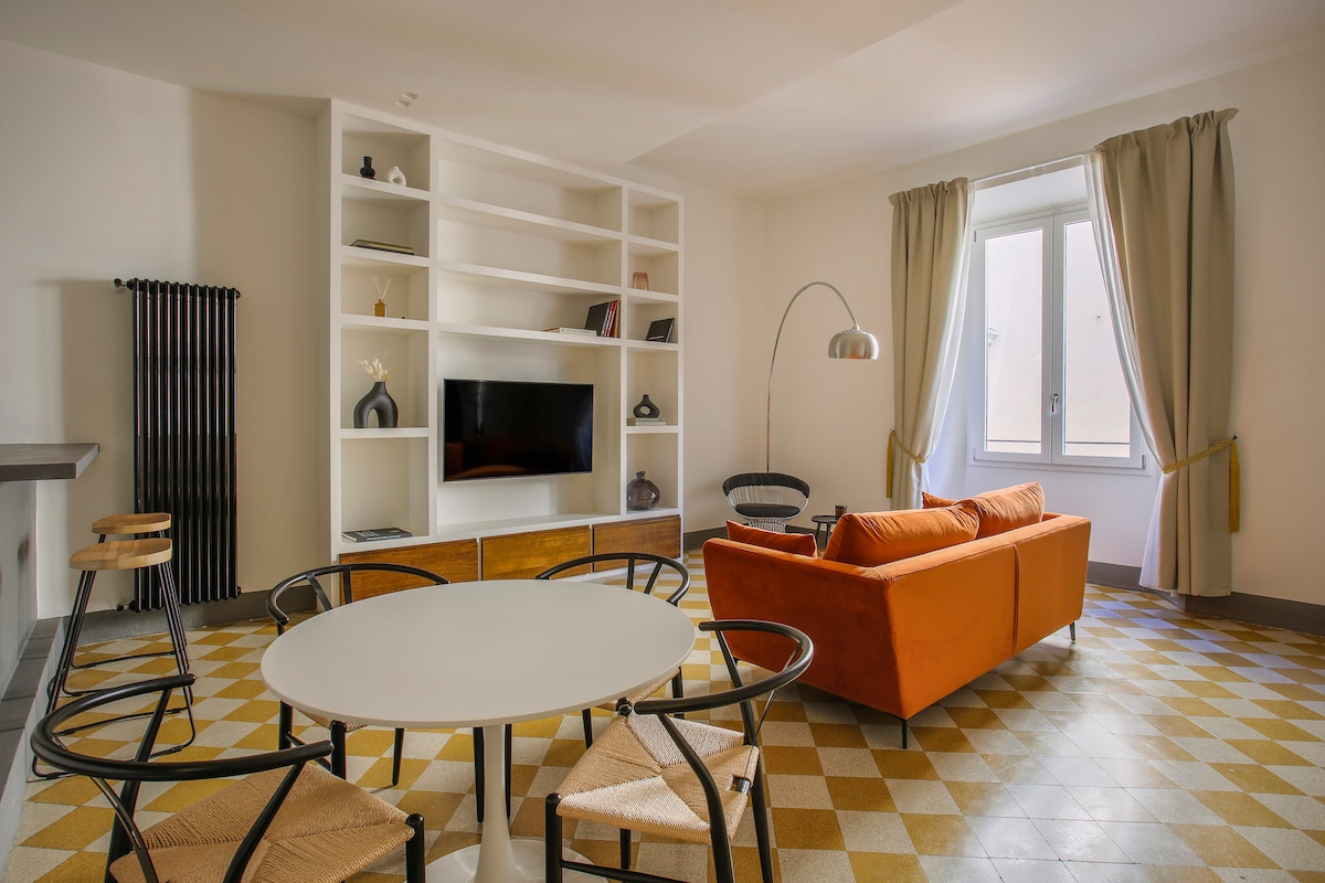 An inviting living area features an orange convertible sofa adjacent to a round dining table with black chairs. A wall-mounted TV is displayed on a white shelf, while natural light enters through a window, complemented by light-colored curtains. Original patterned flooring enhances the space.