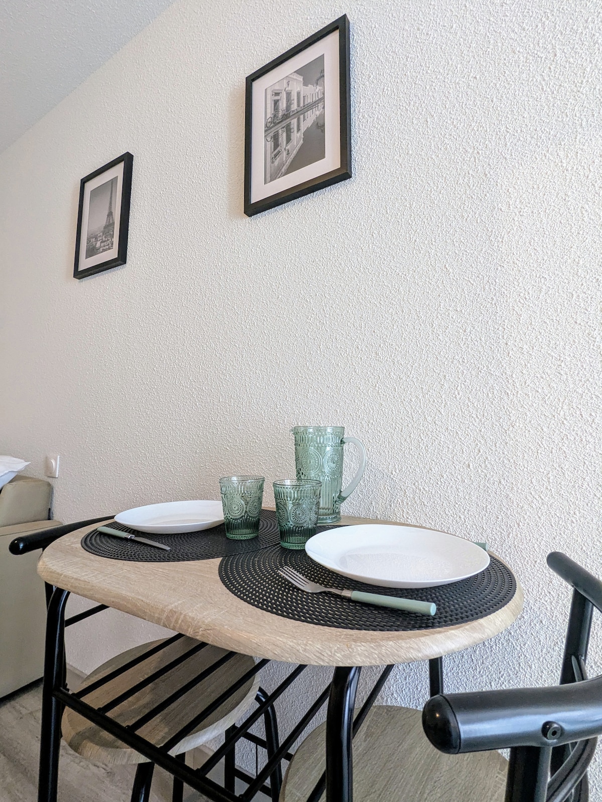 A small dining area is set with two plates and glasses on a textured black placemat. A wooden table is paired with a metal chair. Black and white photographs hang on the wall behind, adding a touch of decor to the space.