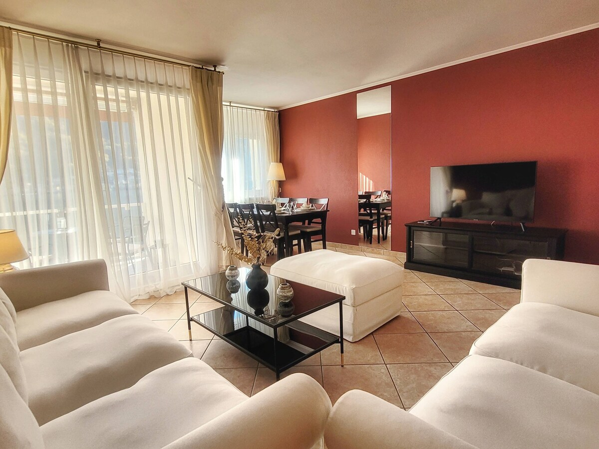 A spacious living area is furnished with a comfortable white sofa and a matching ottoman, complemented by a glass coffee table. Natural light filters through sheer curtains, illuminating the room. In the background, a dining table is visible, set against a warm red wall.