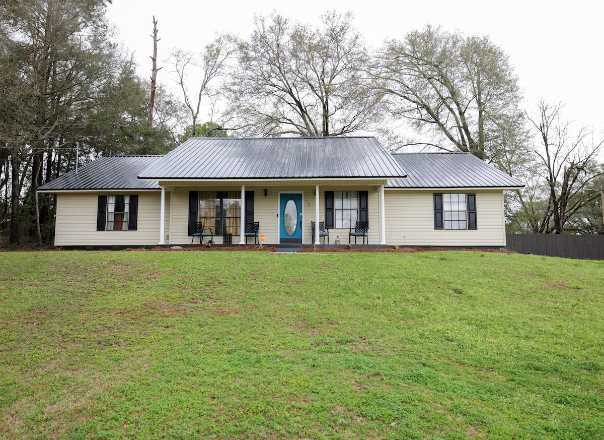 The single-story home is set on a gently sloping lawn, surrounded by mature trees. A welcoming front porch features outdoor seating, while the entrance is highlighted by a blue front door. Side windows provide natural light, complementing the house's neutral exterior.