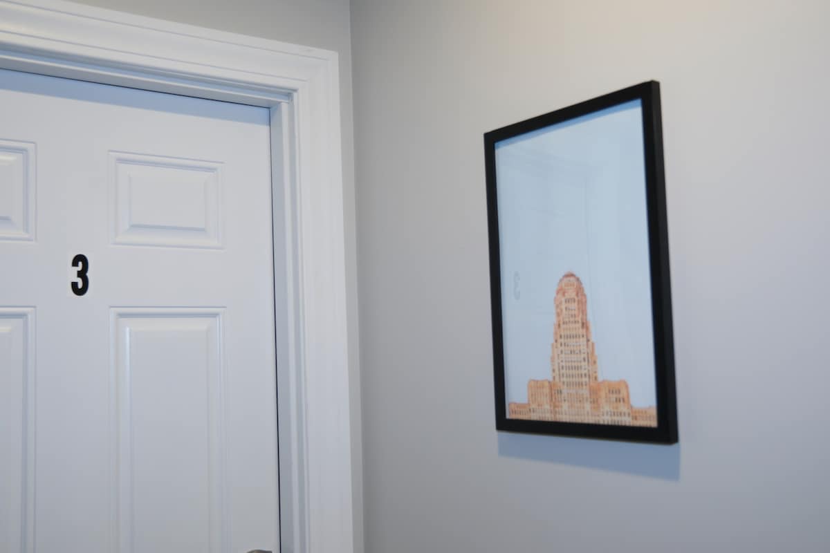 A hallway is depicted with a white door marked with the number three. Adjacent to the door, a framed artwork showcases a stylized building design in warm tones, adding a modern touch to the neutral gray walls.