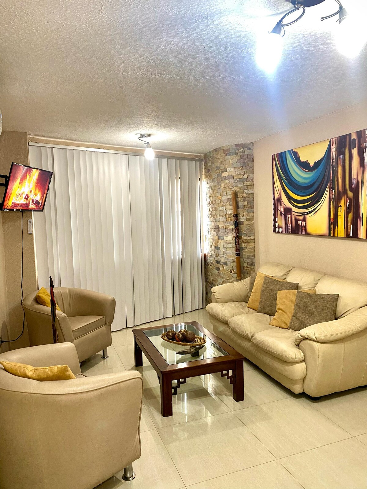 A bright living area is furnished with a cream-colored sofa and two armchairs arranged around a wooden coffee table. A decorative piece is displayed on the table, and a wall-mounted television is visible. Colorful artwork adorns one wall, contributing to a welcoming atmosphere.