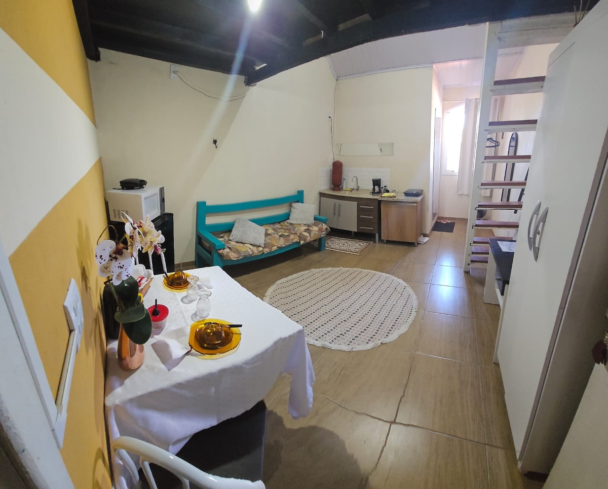 A living space is presented with a cozy seating area featuring a blue couch, a small table set with dishes and glasses, and a circular area rug. A kitchenette is visible in the background, while a staircase leads to an upper level. Natural light enters from a nearby window.