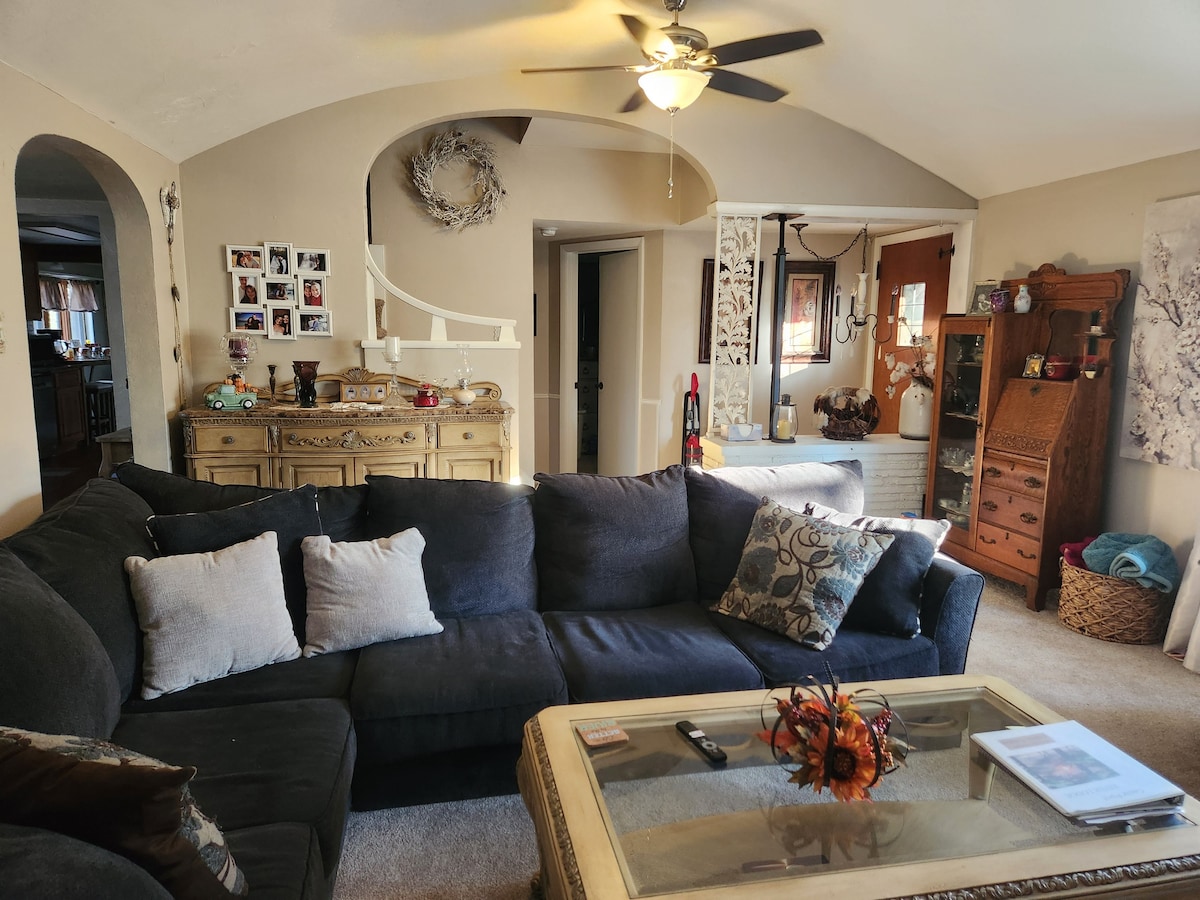 A spacious living area features a dark sectional sofa adorned with decorative pillows. A glass coffee table sits in the center, complemented by a floral centerpiece. The background includes a wooden sideboard and various decorative accents, illuminated by natural light from a nearby window.