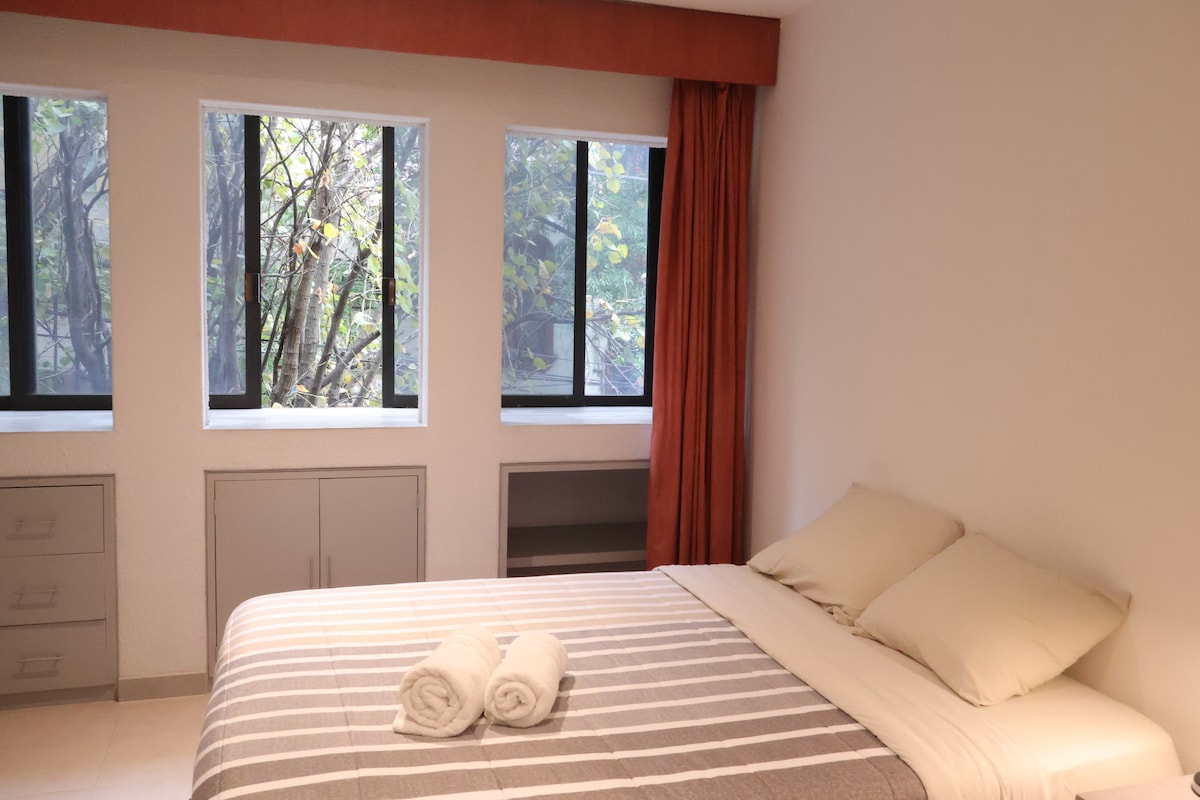 A well-lit bedroom features a spacious bed with soft, neutral linens. Natural greenery is visible through the three large windows, framed by orange curtains. Minimalist cabinetry is present, enhancing the clean and uncluttered look of the space.